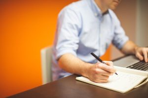 Un homme note des idées en écrivant, avec ordinateur et carnet